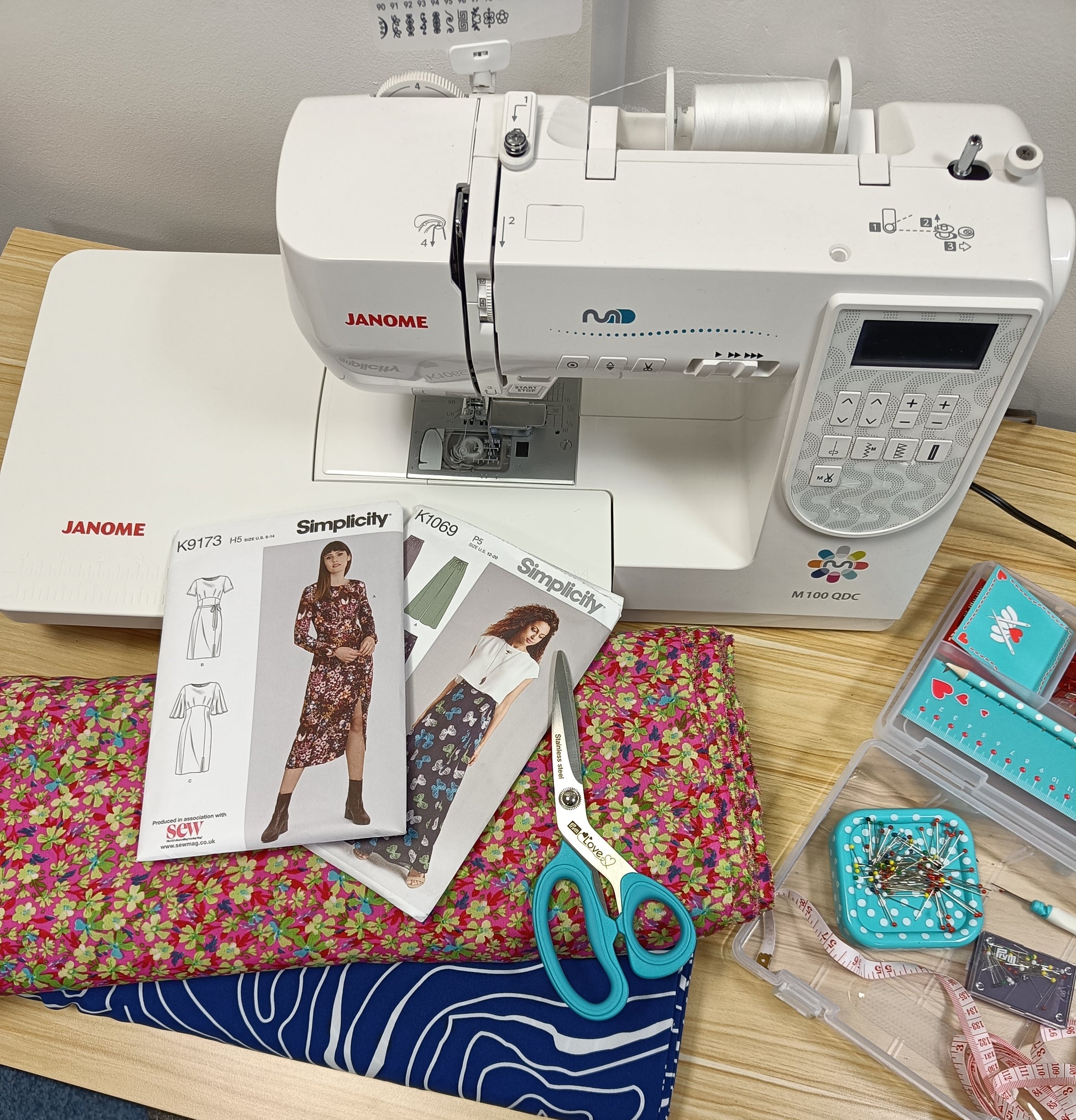 Janome sewing machine with 2 patterns for ladies wear, a pair of turquoise scissors and two rolls of patterned fabric